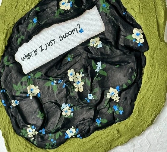 A textured relief piece on a moss green ground with the words 'What if I just bloom?'